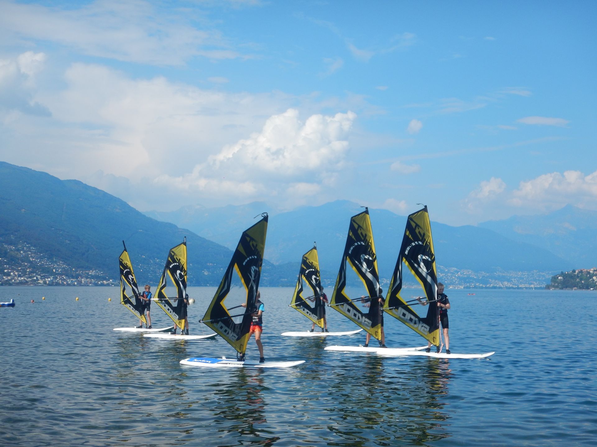 Windsurfers with black-and-yellow sails gliding across a calm blue lake with mountains in the background