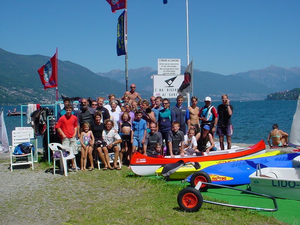 Group posing by sailboats on a sunny lakeshore with mountains in the background