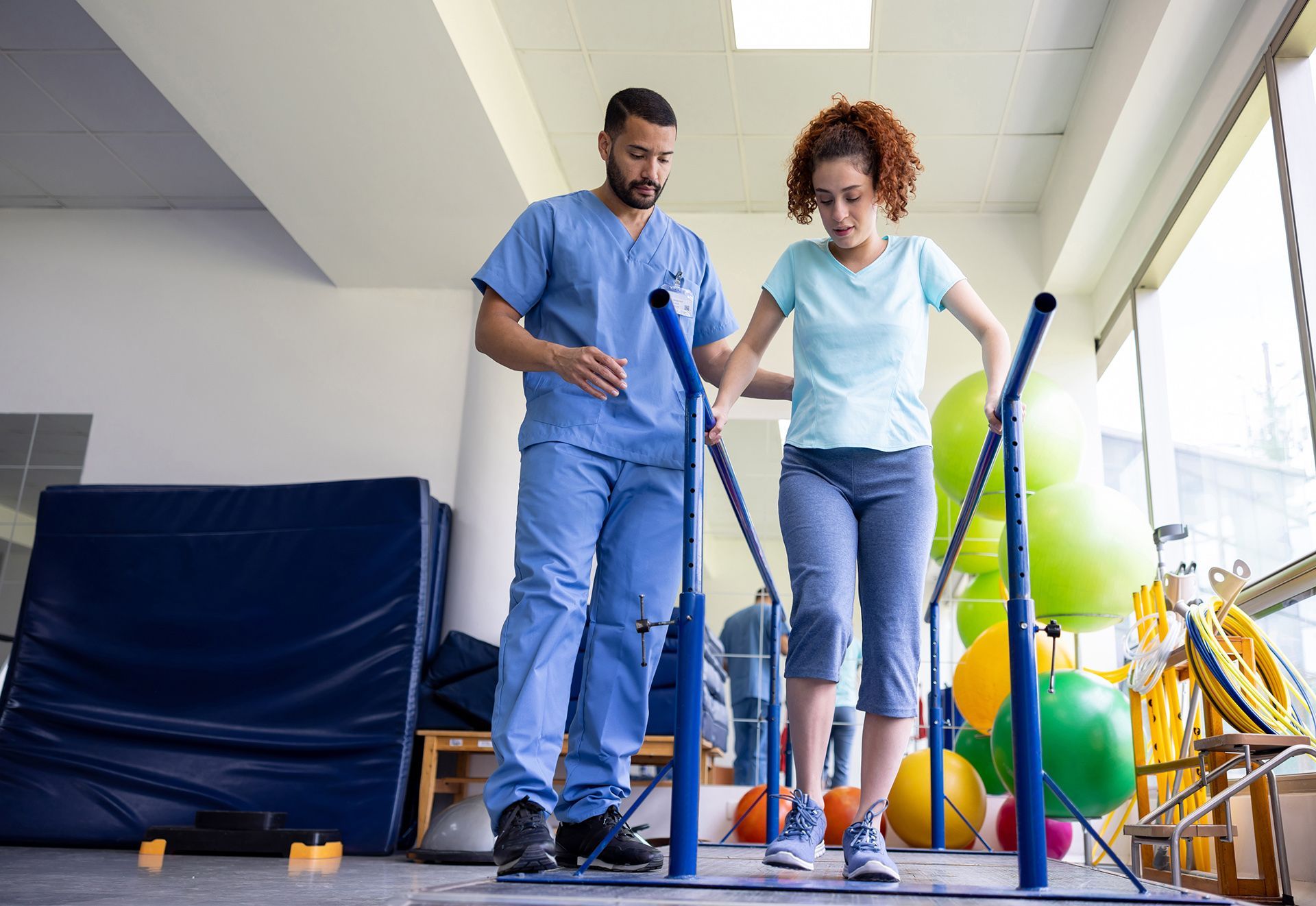 Un hombre ayuda a una mujer a caminar sobre barras paralelas en un gimnasio.