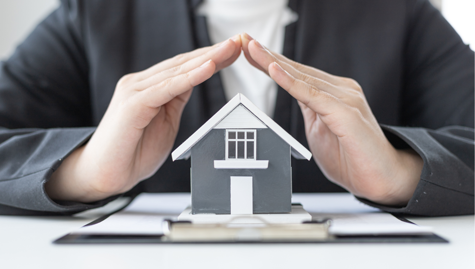 Hands sheltering a small house model on a desk, symbolizing home protection and insurance