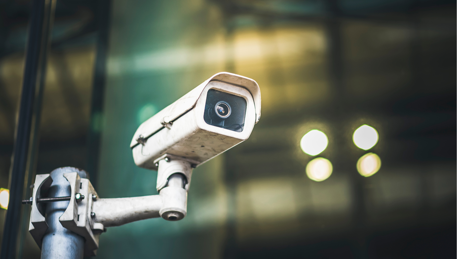 White security camera mounted on a pole, overlooking a blurred indoor area with lights