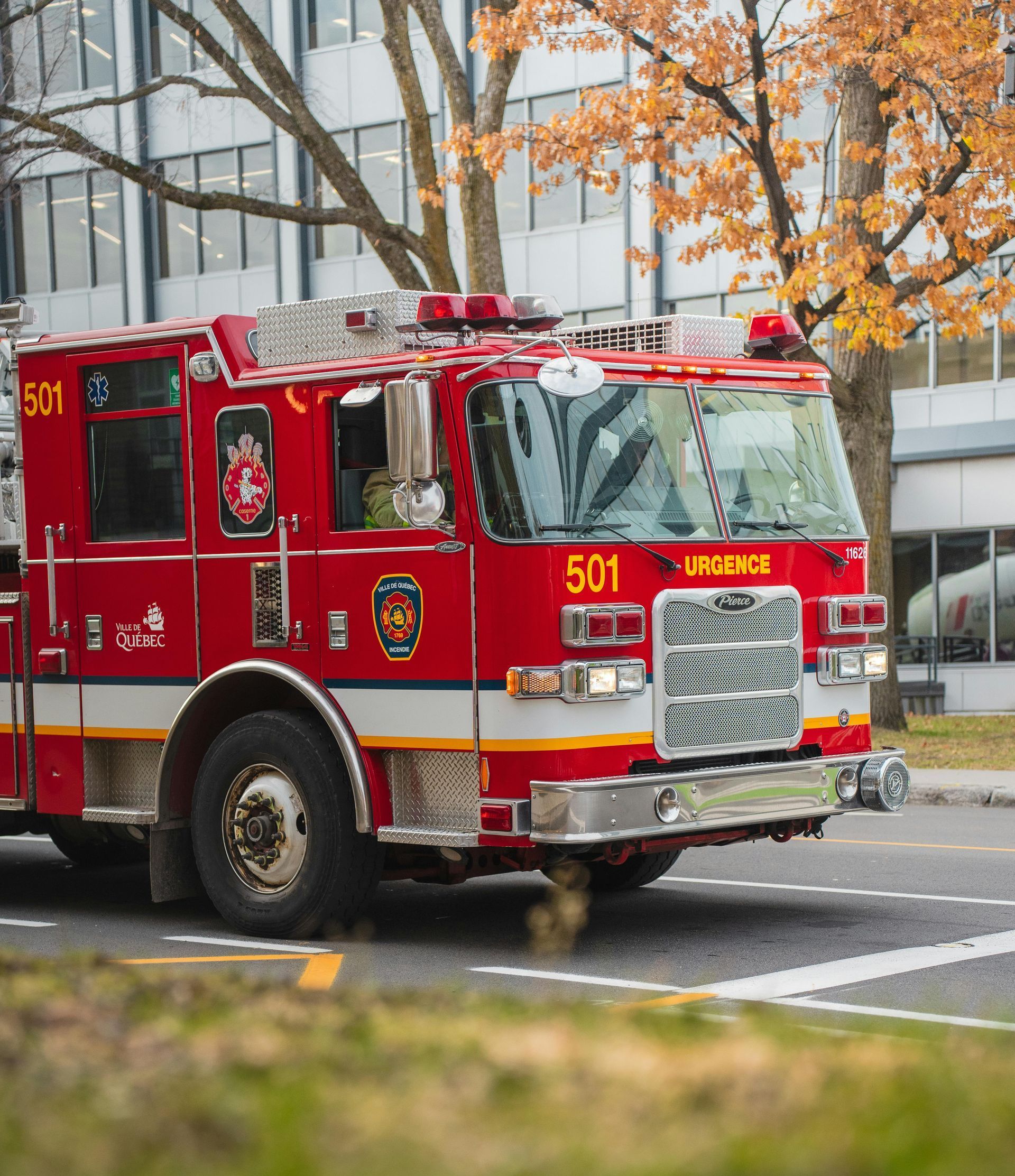 A red fire truck numbered 501 drives along a paved road near a building with autumn trees in the background.
