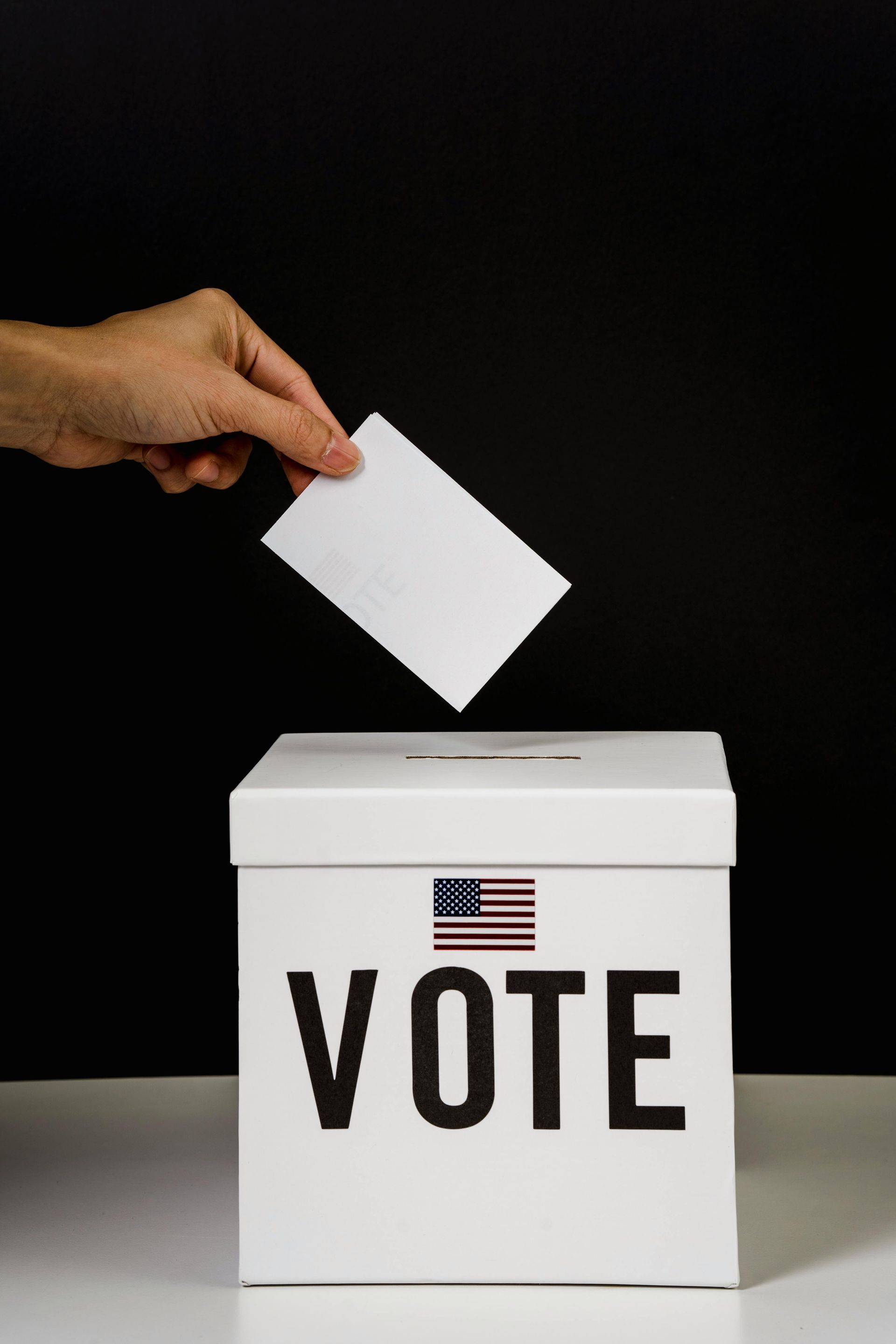 A hand places a white ballot into a white box labeled