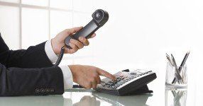 A person in a dark suit holding a telephone handset to their ear while dialing a desk phone.