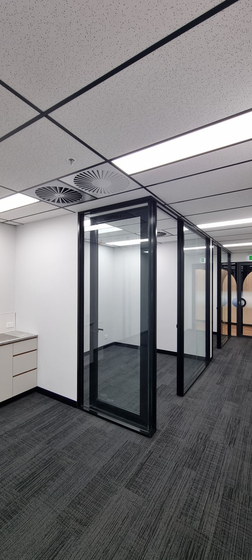 An office interior with white desks, black computer monitors, and chairs on a wood floor under long hanging lights.