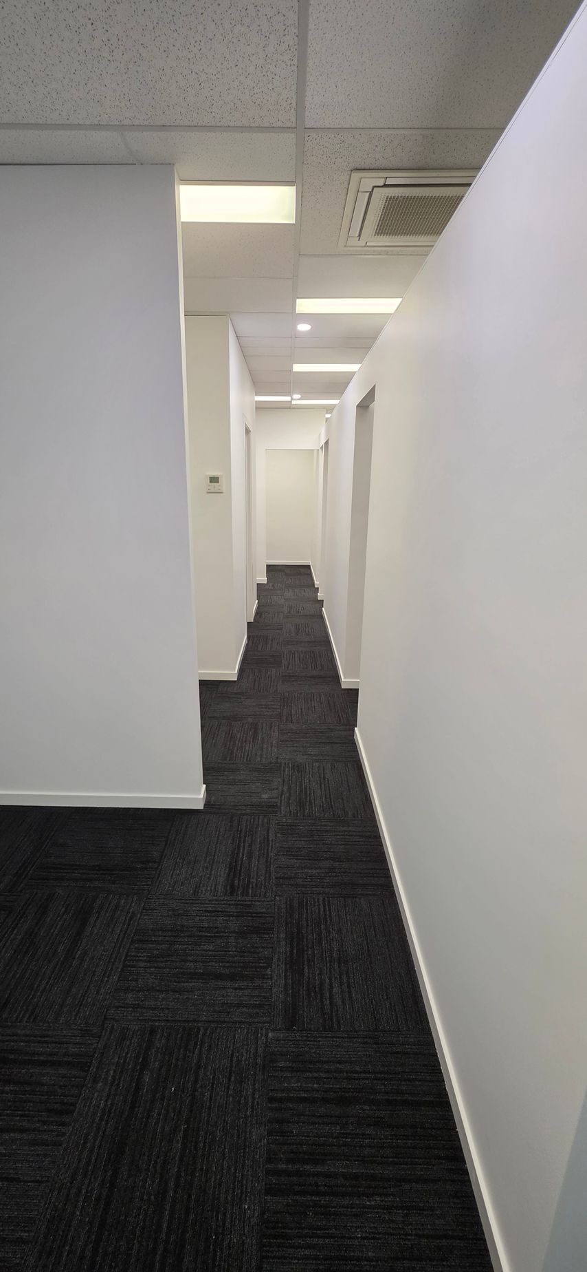 A bright, empty office space with rows of modular beige cubicles and desks leading toward a hallway and doorway.