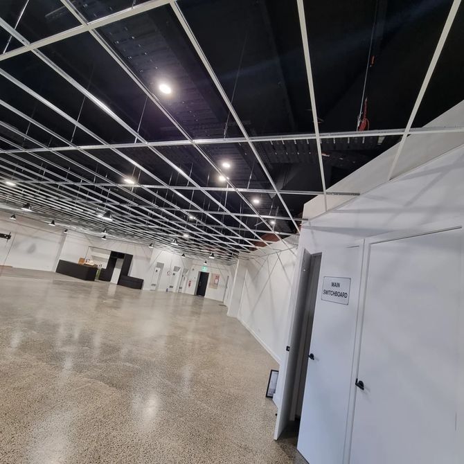 A large, empty hall with a concrete floor, a black ceiling, and an unfinished white metal grid frame hanging above.