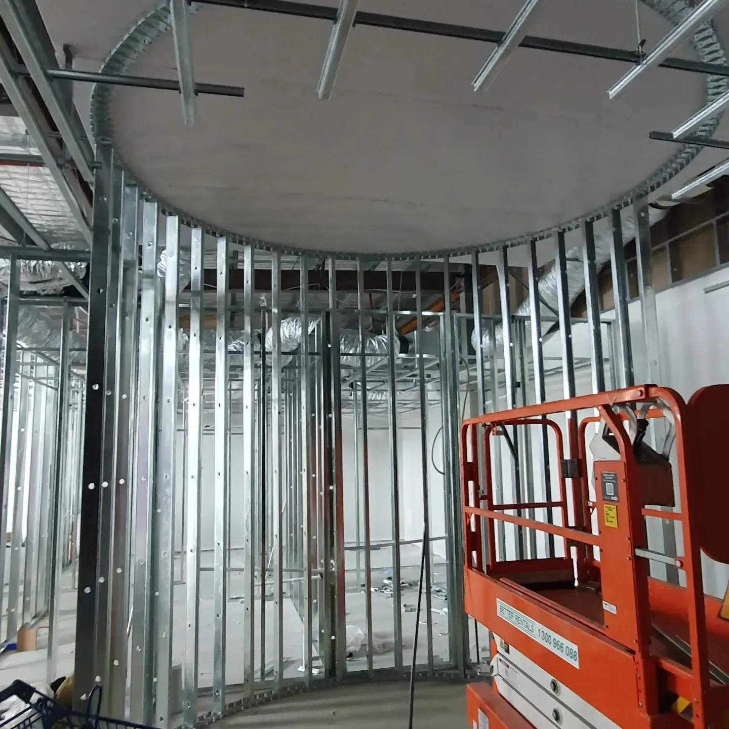 Construction site showing a circular metal stud wall frame with a curved ceiling track and an orange scissor lift.