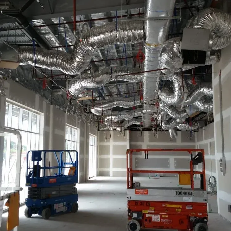 An indoor construction site features an exposed ceiling with silver ductwork, with blue and orange scissor lifts below. An indoor construction site features an exposed ceiling with silver ductwork, with blue and orange scissor lifts below.