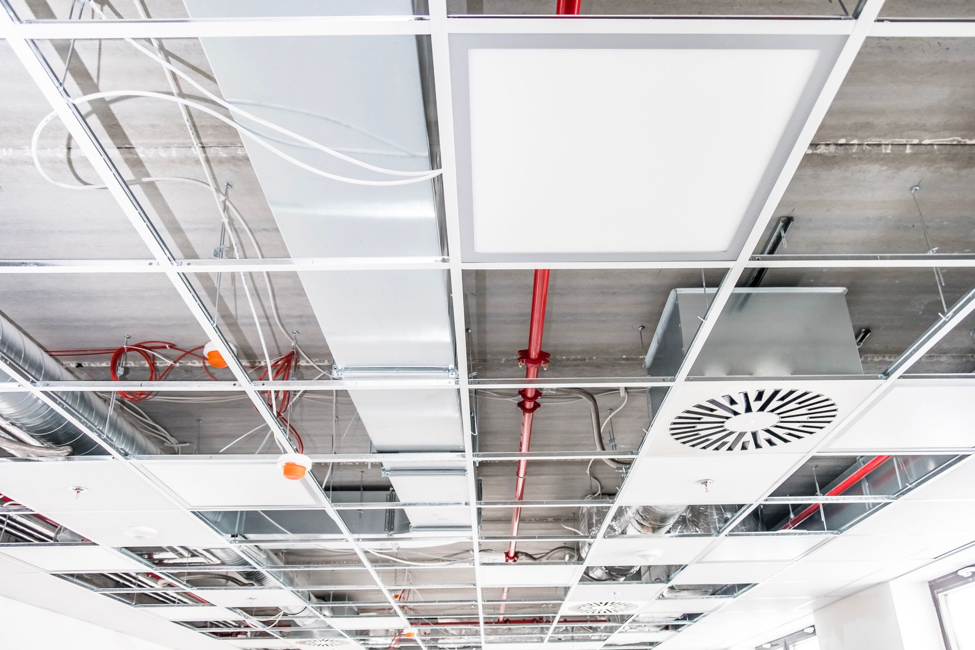 A drop ceiling under construction, featuring a white light panel, a circular ventilation grille, and exposed wiring.