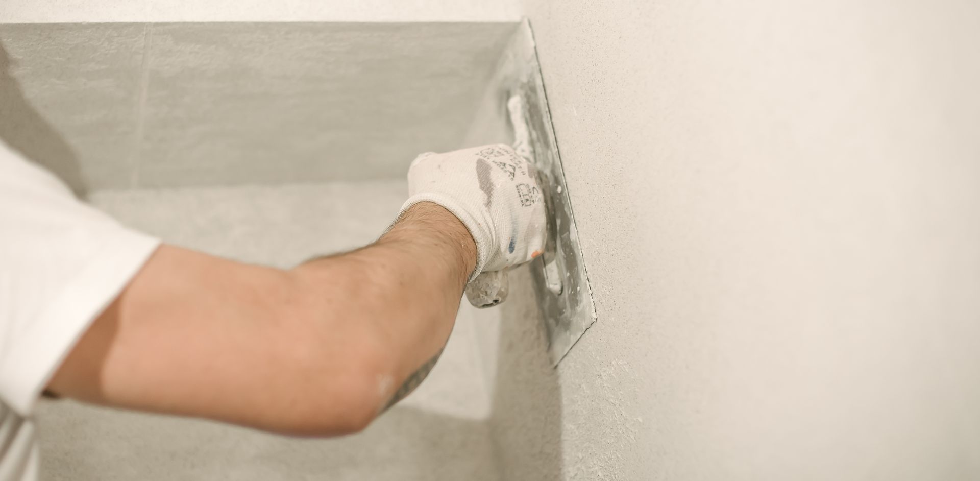 A person wearing a white glove uses a metal trowel to smooth plaster onto a light-colored wall. A person wearing a white glove uses a metal trowel to smooth plaster onto a light-colored wall.