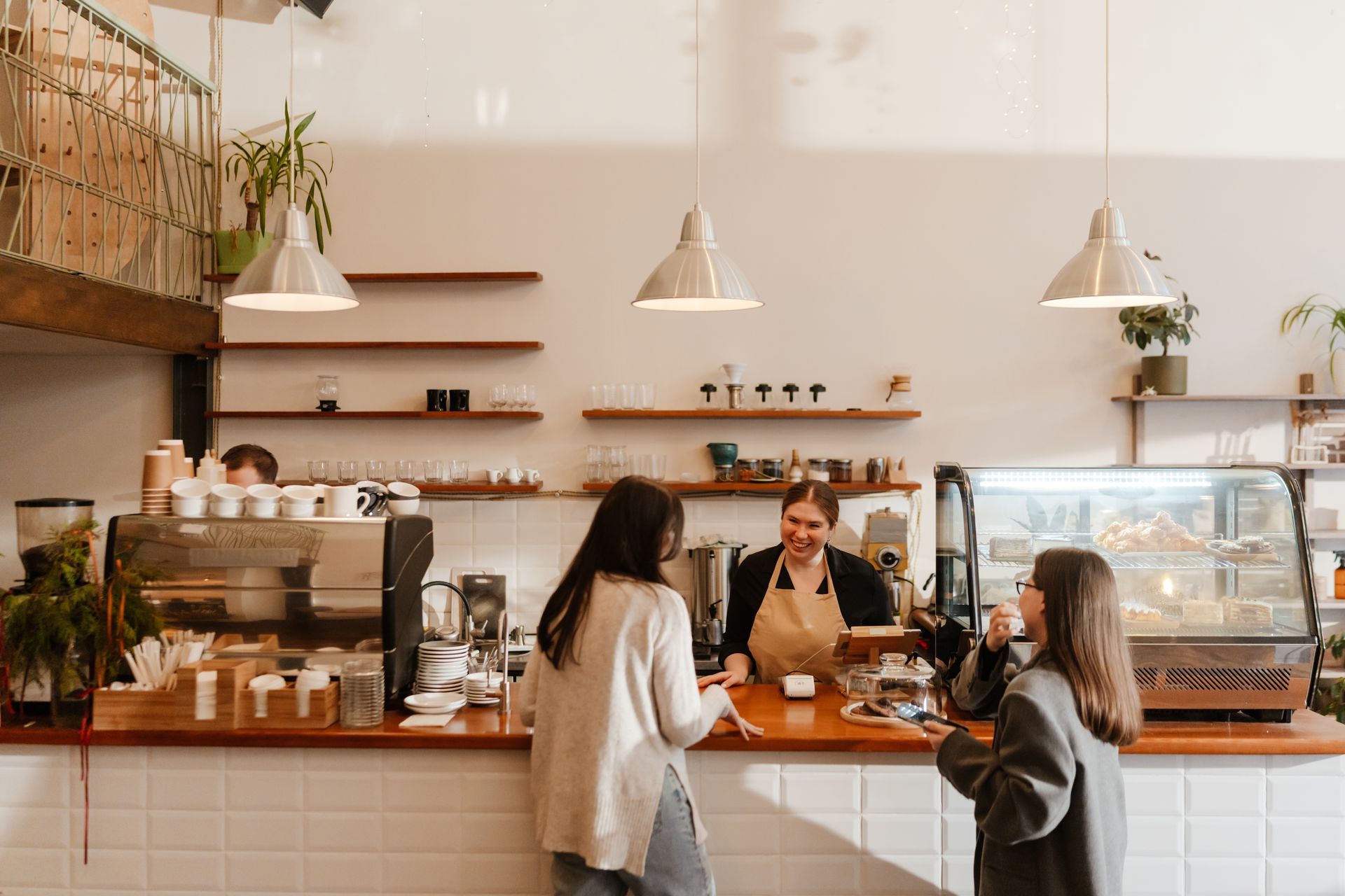 A barista behind a cafe counter takes an order from two customers in a brightly lit, modern coffee shop.