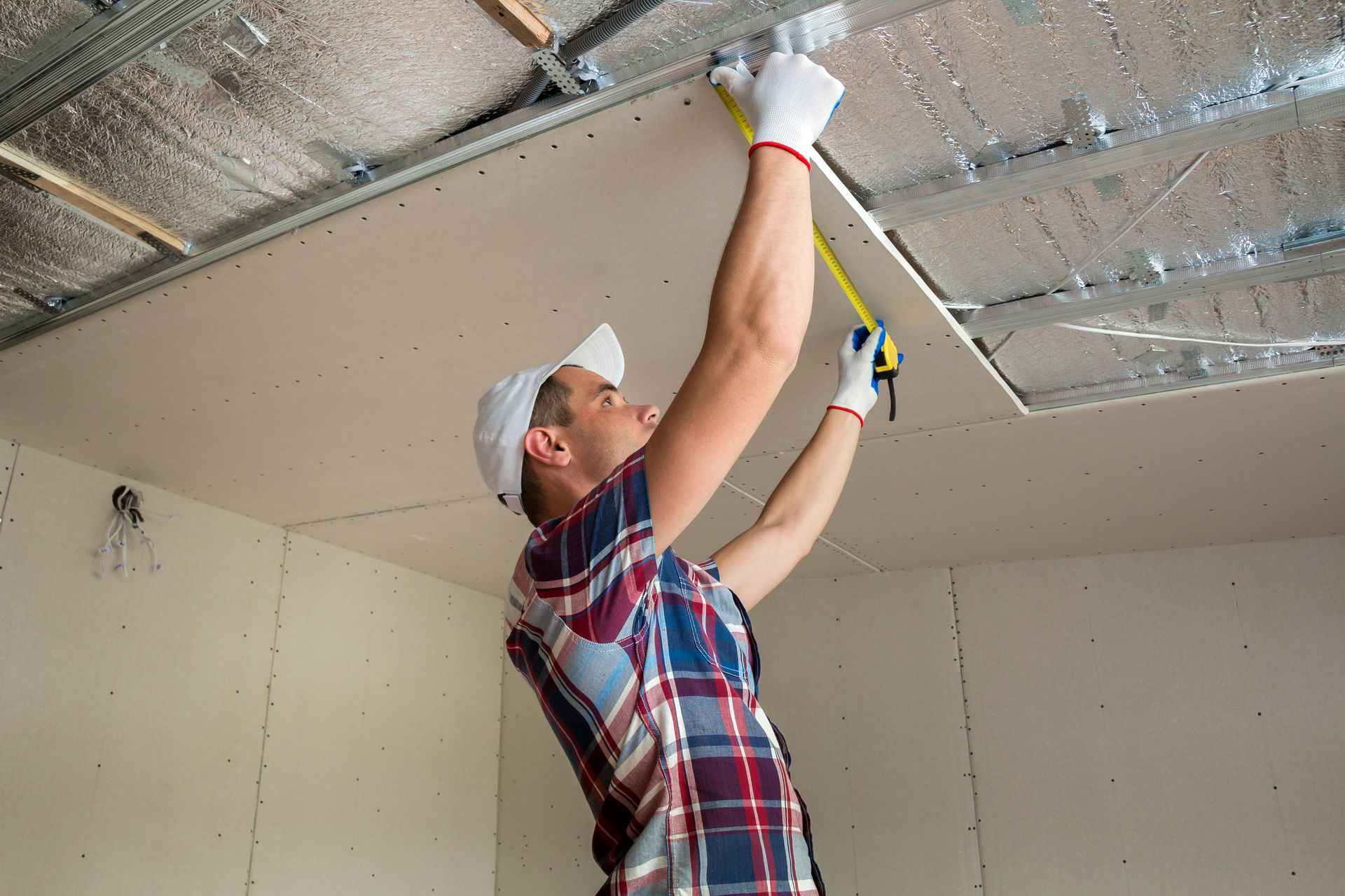A person in a plaid shirt and white cap uses a yellow measuring tape to measure a ceiling during a renovation.