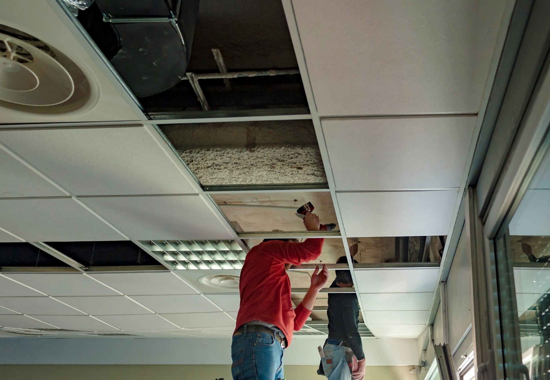 Two people working on a suspended ceiling in an office, installing or repairing panels in the metal framework.
