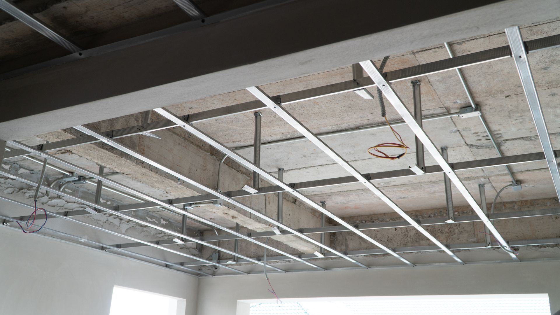 A metal grid frame for a suspended ceiling installed below a concrete ceiling with exposed wiring in a room.