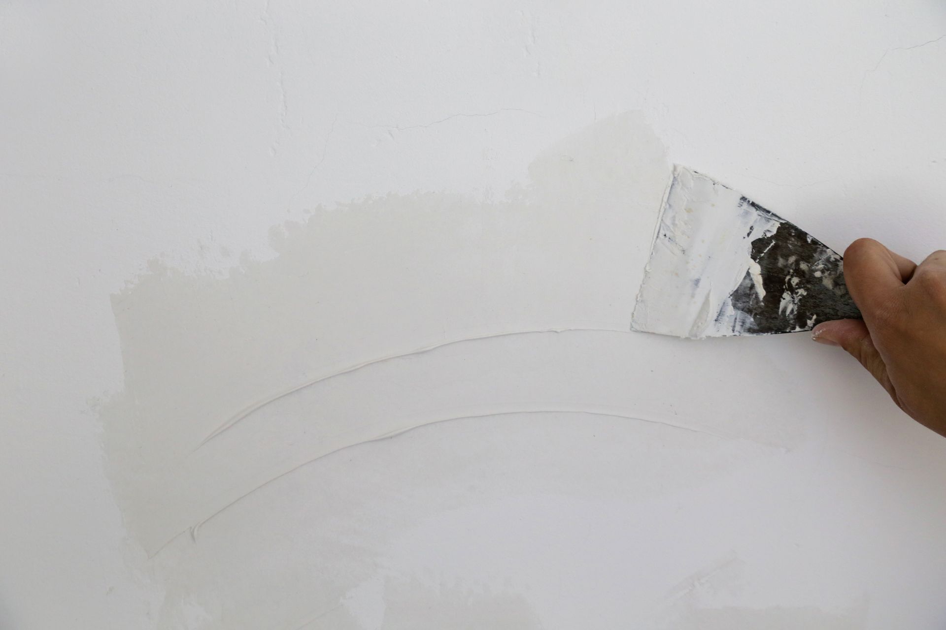 A hand uses a metal putty knife to apply white spackle or filler to a smooth white wall.