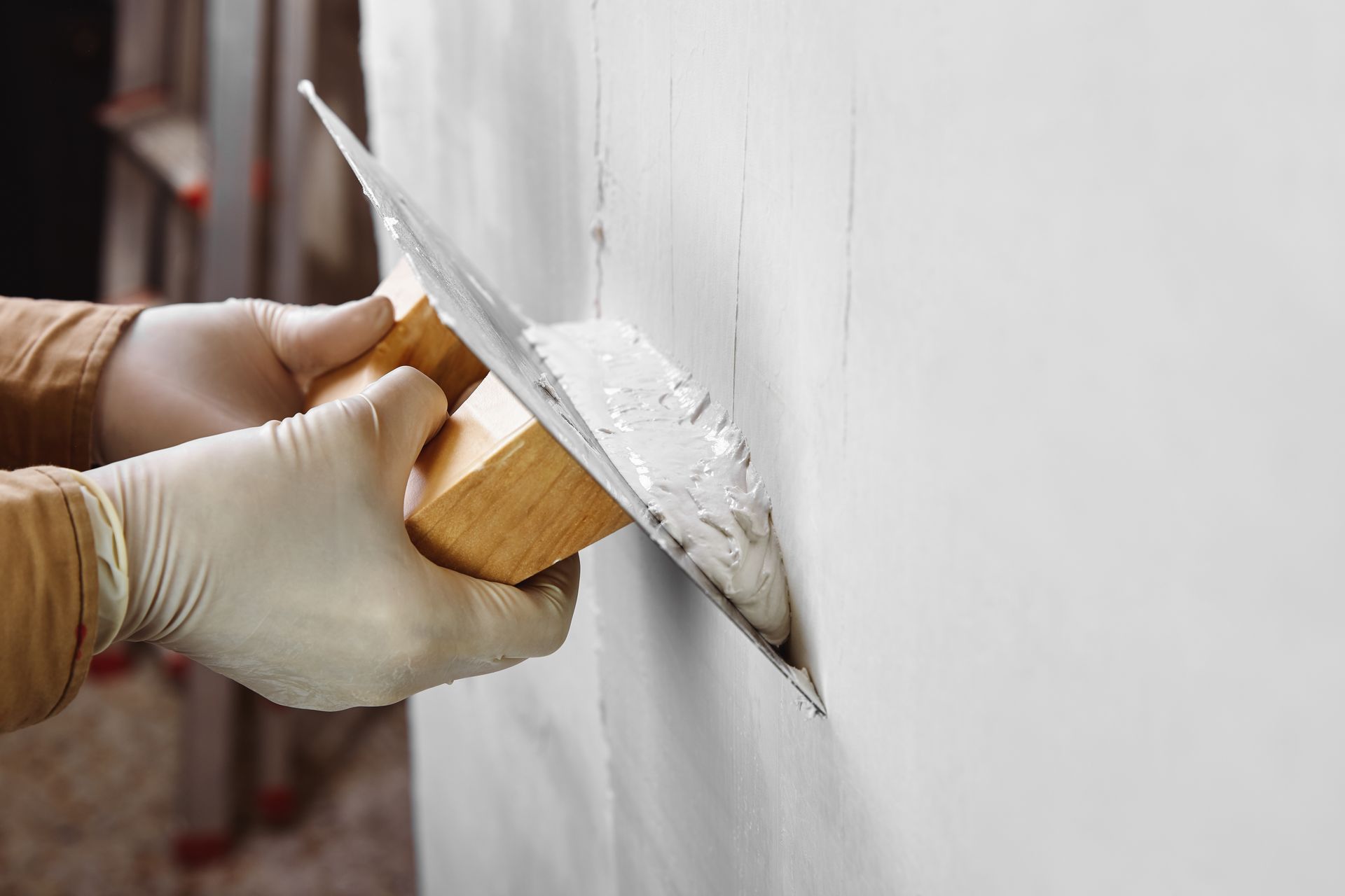 A gloved hand uses a metal trowel to smooth white joint compound onto a wall. A gloved hand uses a metal trowel to smooth white joint compound onto a wall.