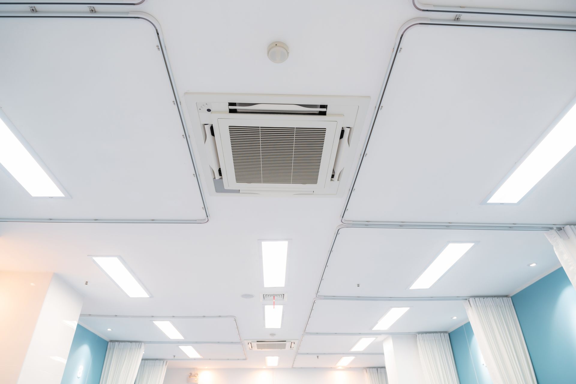 A view of a modern ceiling with recessed lighting panels, a central air conditioning unit, and white privacy curtains.