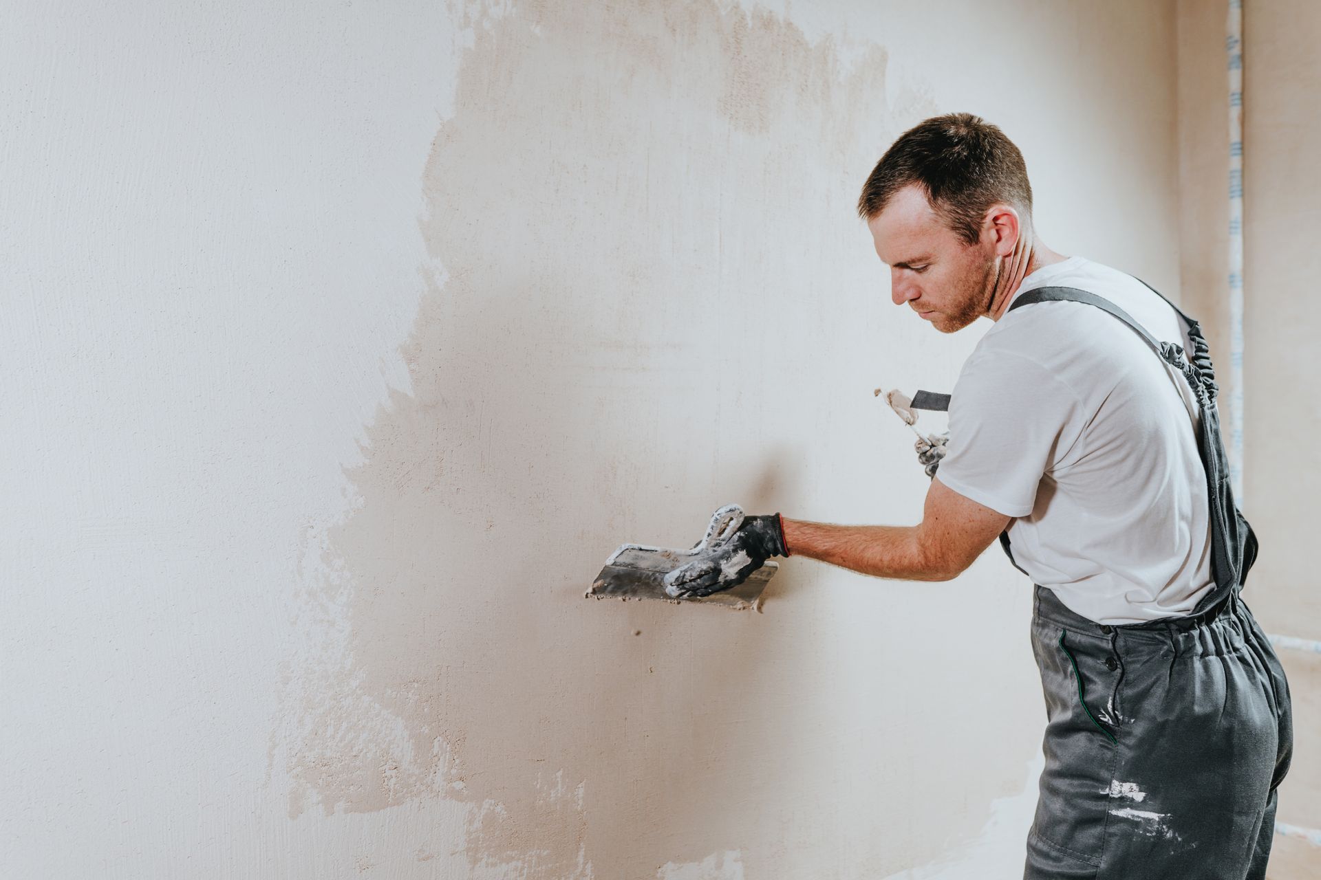 A worker in grey overalls applies plaster to a wall with a metal trowel.