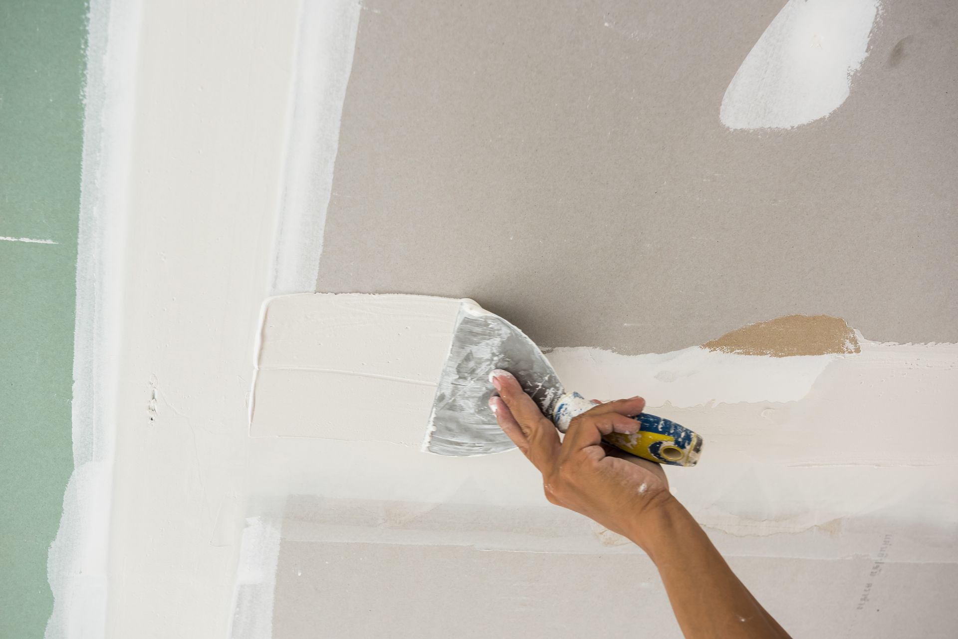 A hand uses a metal putty knife to spread joint compound over a seam on a drywall ceiling. A hand uses a metal putty knife to spread joint compound over a seam on a drywall ceiling.