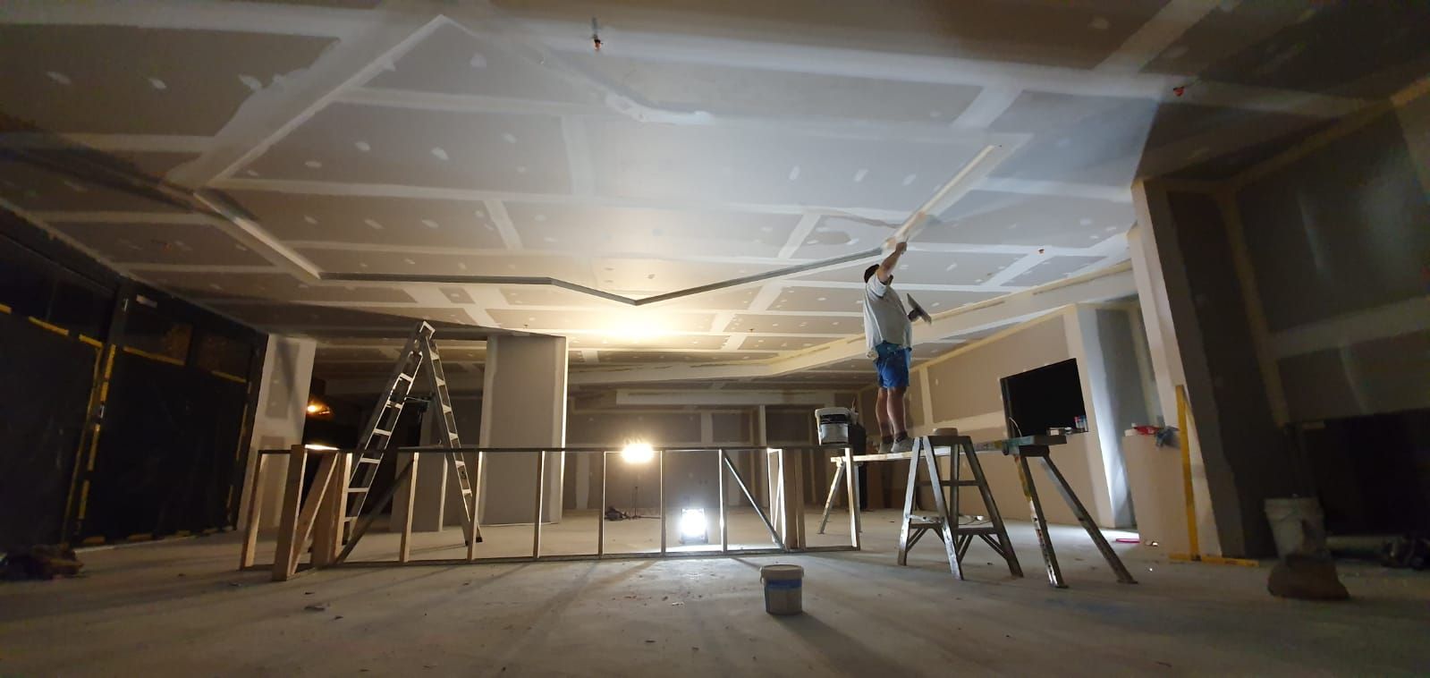 A worker in grey overalls applies plaster to a wall with a metal trowel.