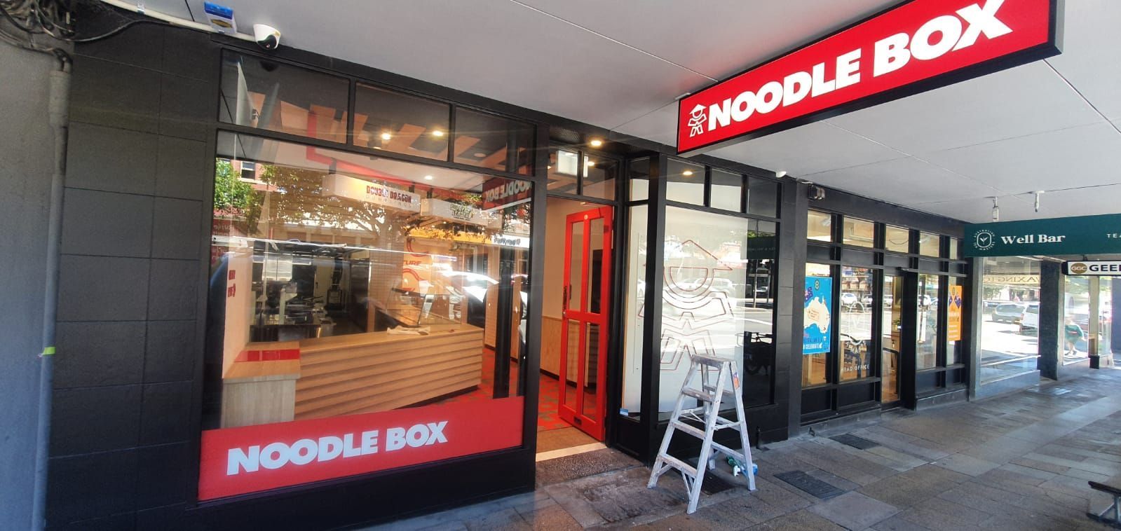 The storefront of a Noodle Box restaurant featuring black walls, large windows, and a bright red sign.