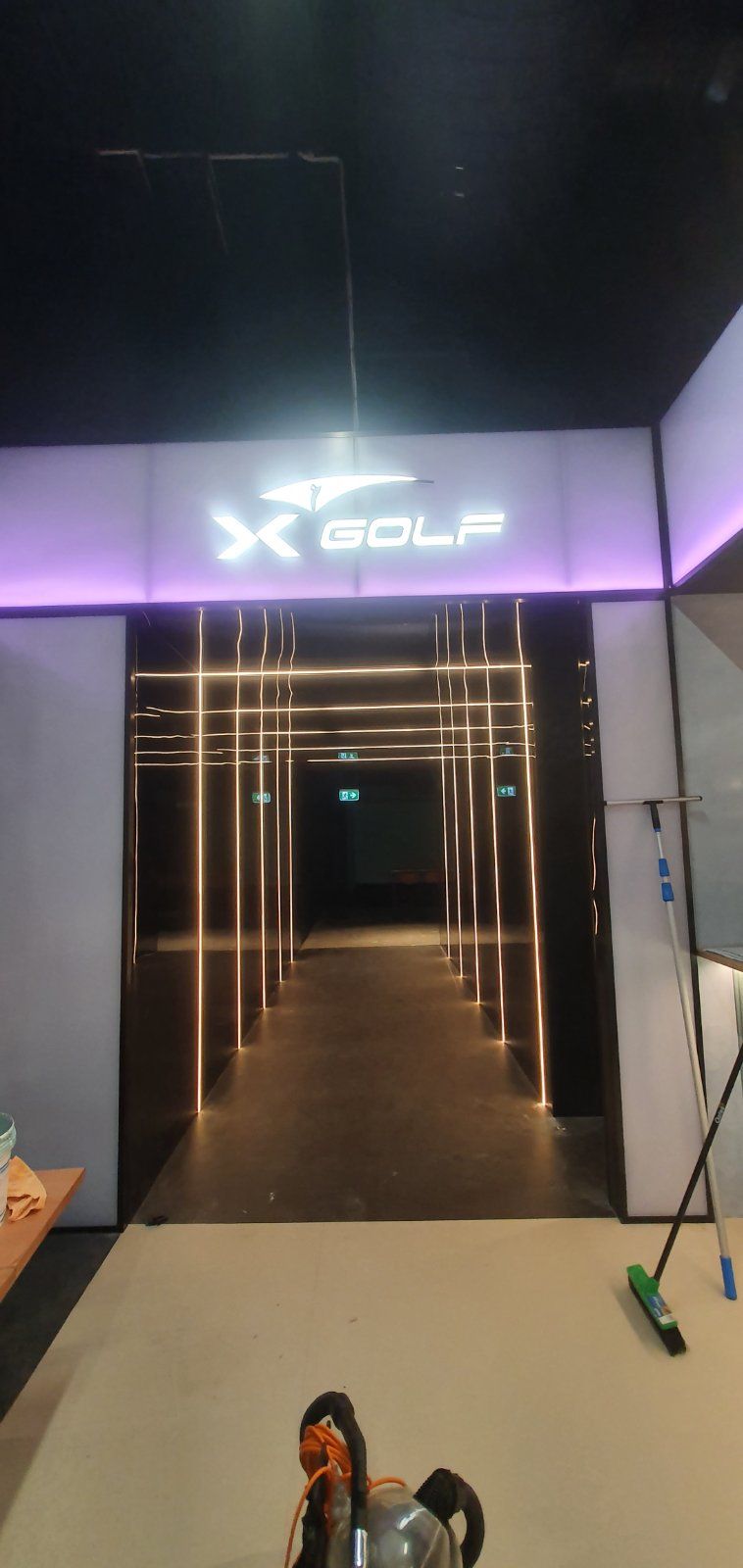Entrance to an X-GOLF venue with a glowing sign above a dark hallway illuminated by thin vertical LED light strips.