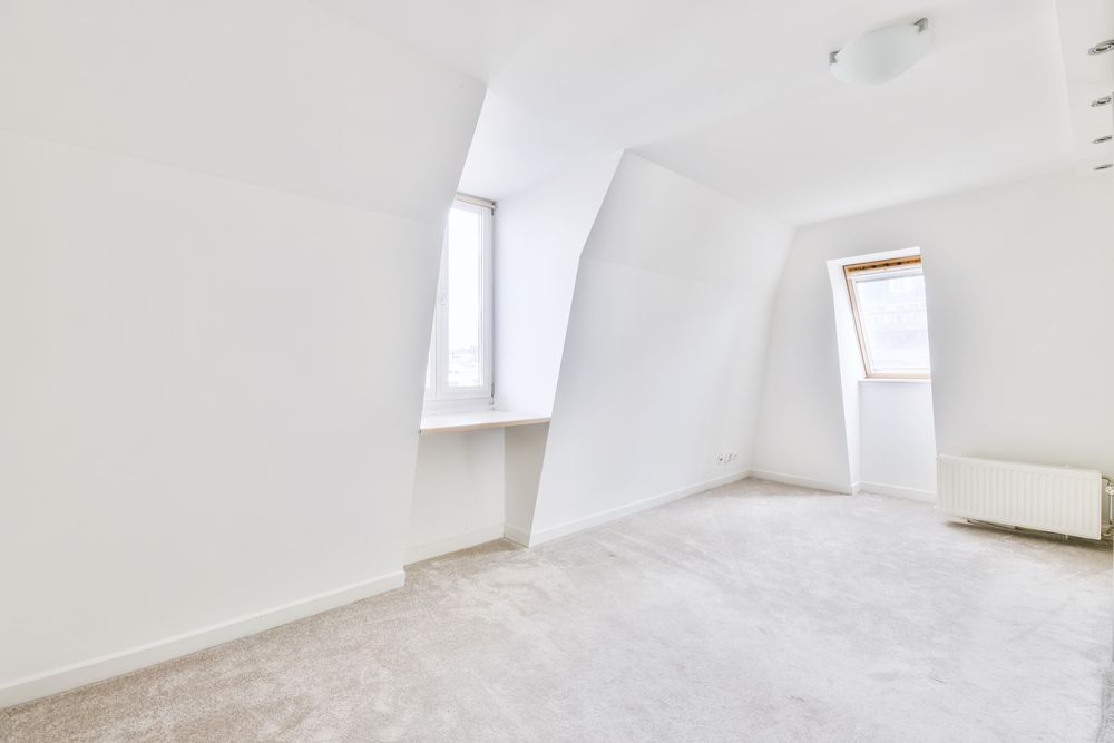 An empty attic room with white walls, slanted ceilings, two dormer windows, and light gray carpeted flooring.