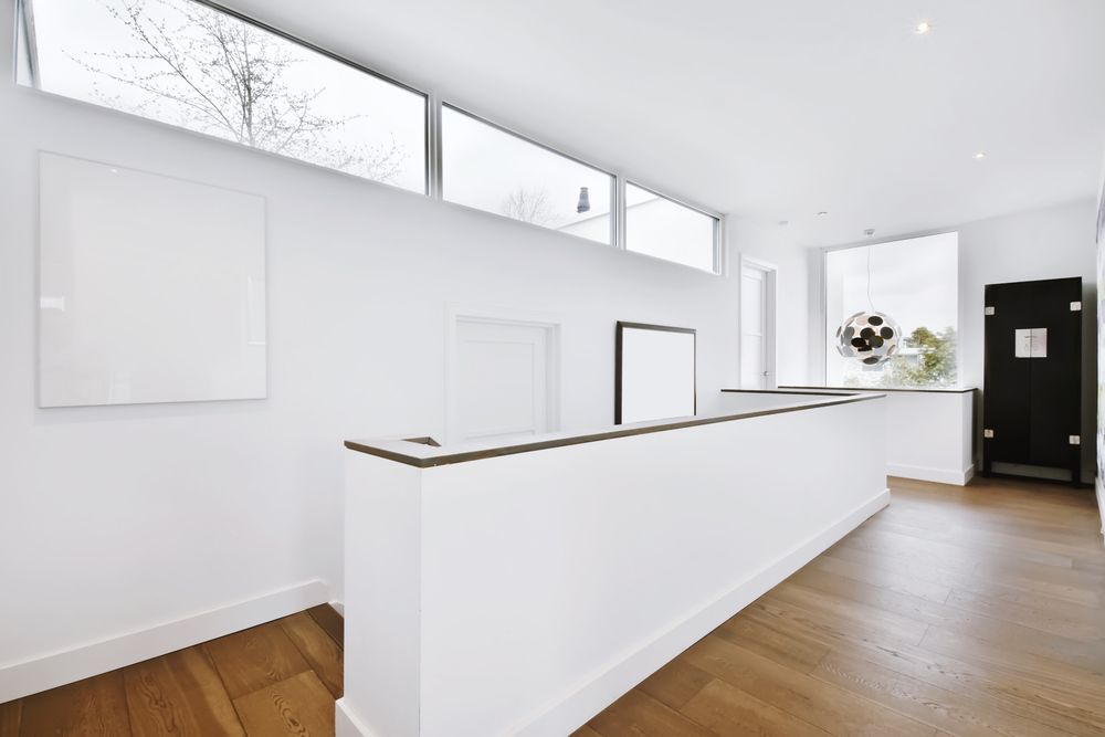 A minimalist, bright hallway with white walls, light wood flooring, a staircase railing, and horizontal clerestory windows.