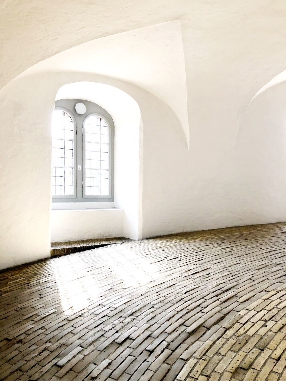 An arched window lets light into a minimalist, white-walled room with a curved brick floor.