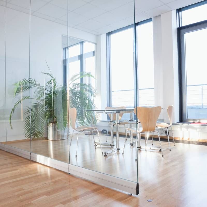 Modern meeting room with a glass wall, wooden floors, a table with four wooden chairs, and a large potted plant.