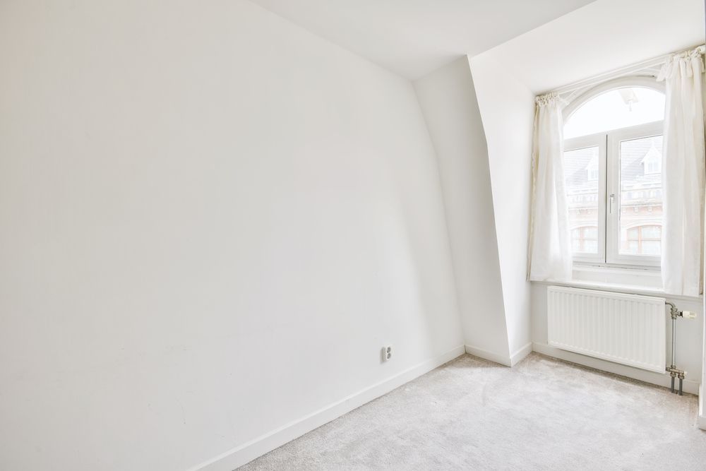 An empty room with white walls, carpeted floors, a window with white curtains, and a wall-mounted radiator.