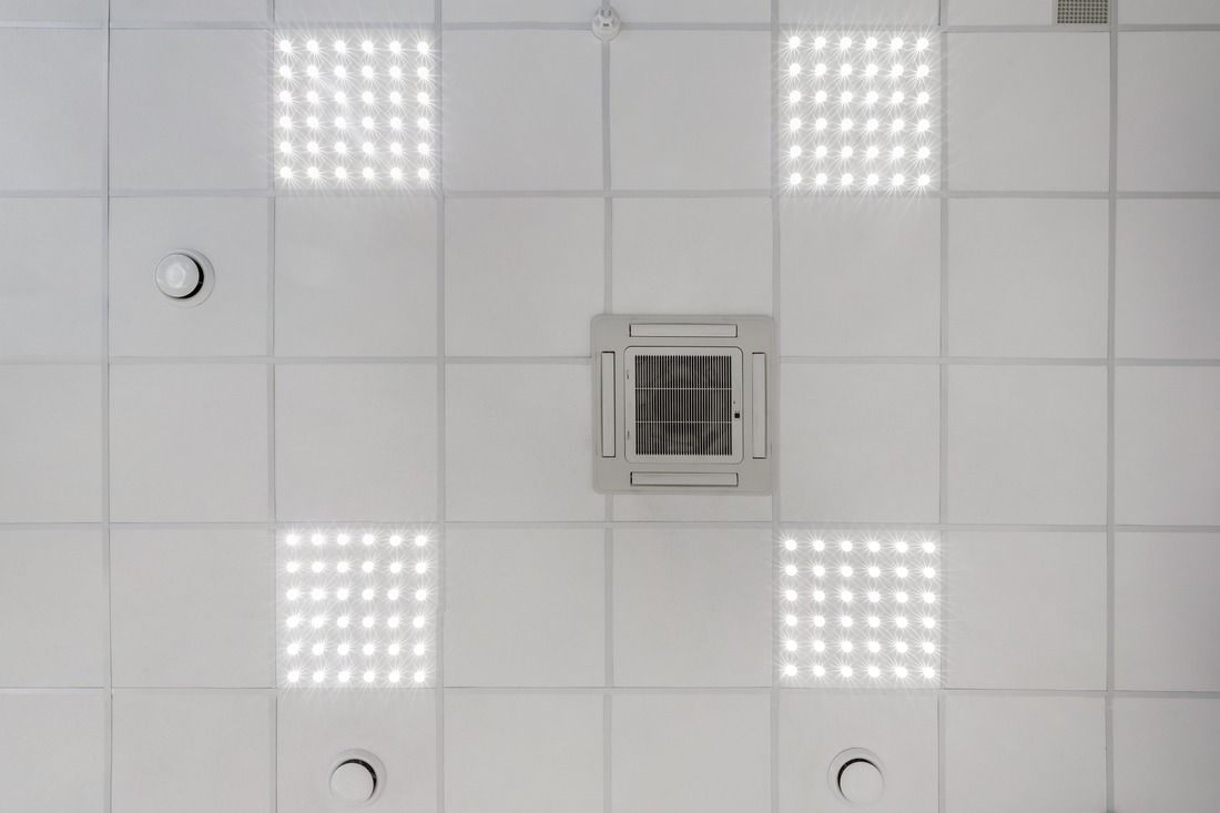 A white drop ceiling with four recessed square LED light panels, a central air conditioning vent, and three smoke sensors.