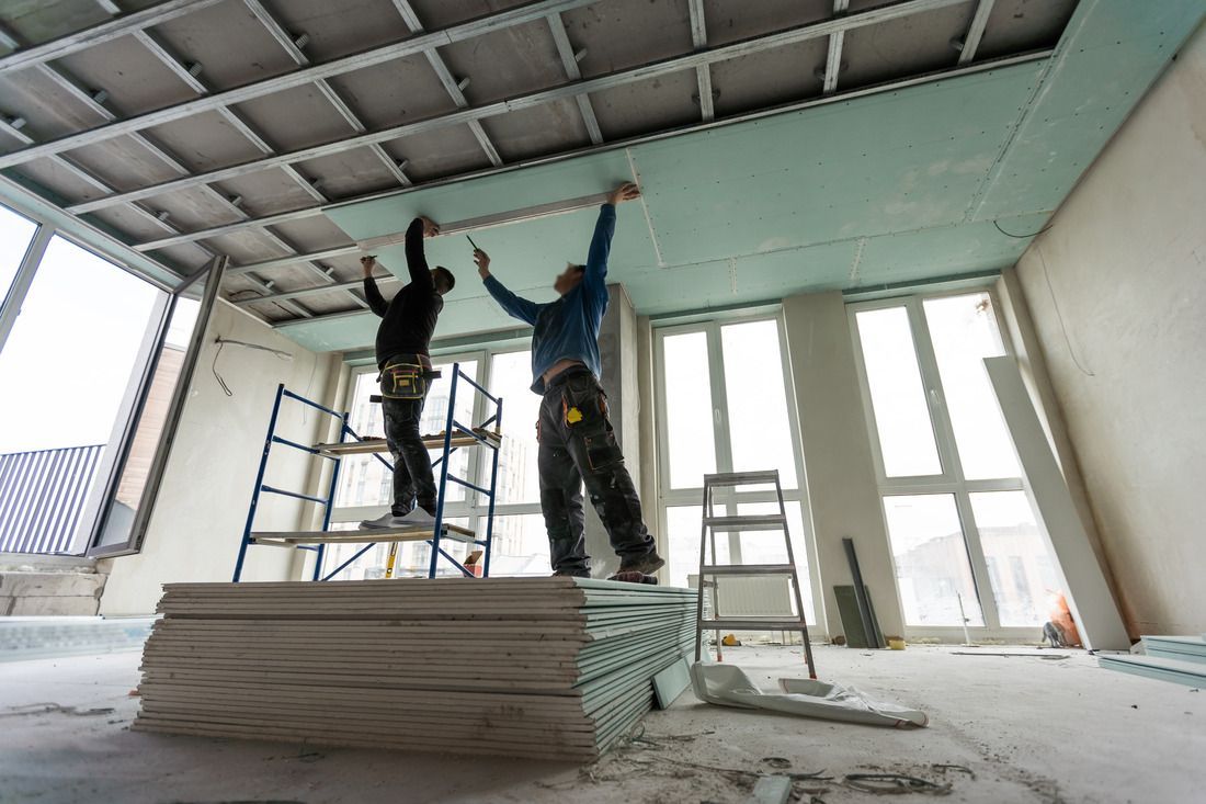 Two workers install drywall panels onto a metal ceiling frame in a room under construction with large windows.