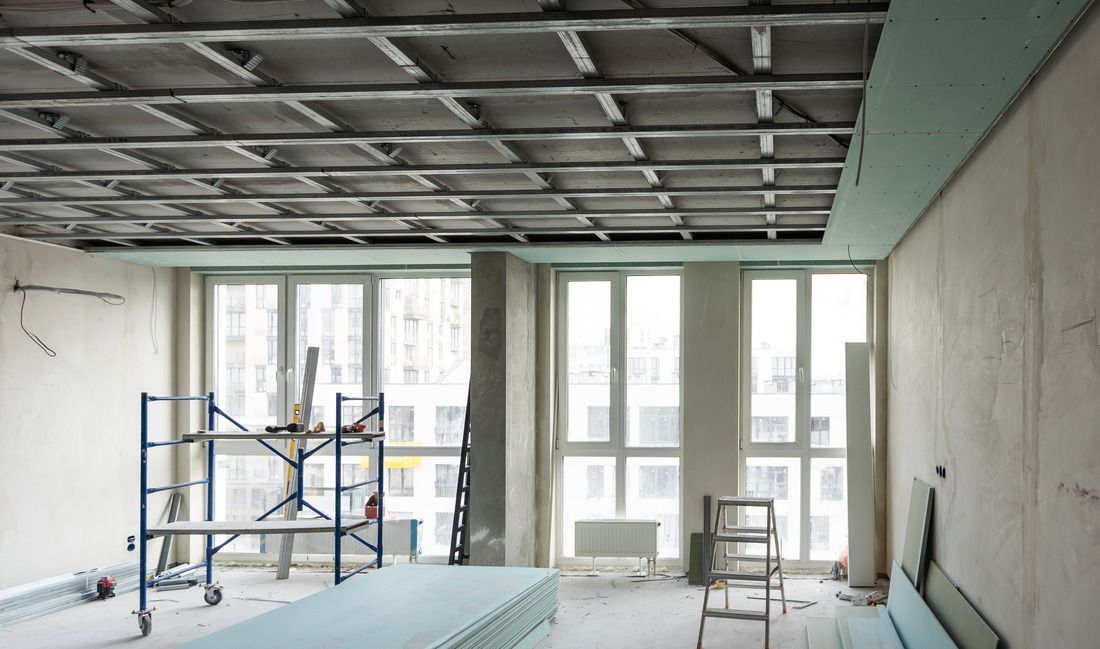 A room under renovation with a metal ceiling grid frame, bare walls, windows, and scaffolding.