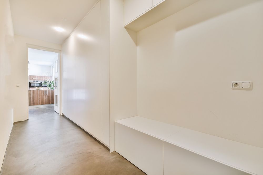 A bright, minimalist hallway with white walls, a built-in bench, and a glimpse into a kitchen with wooden cabinets.