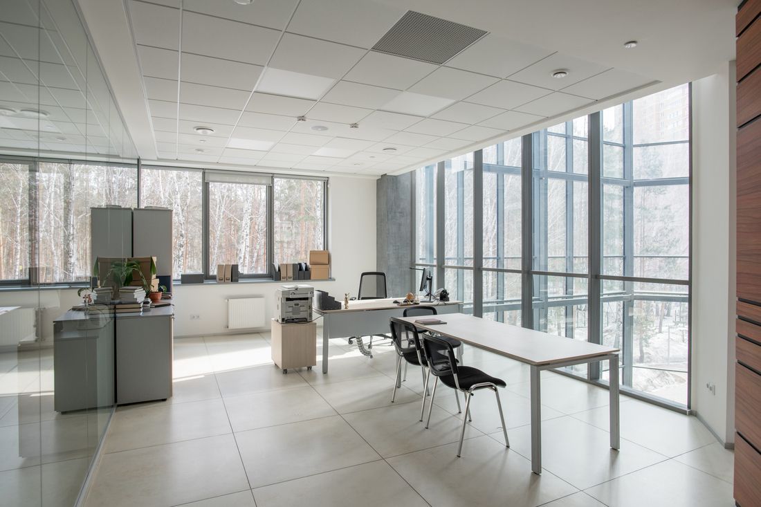 A bright, modern office space featuring large windows, white tile floors, a meeting table with chairs, and a desk.
