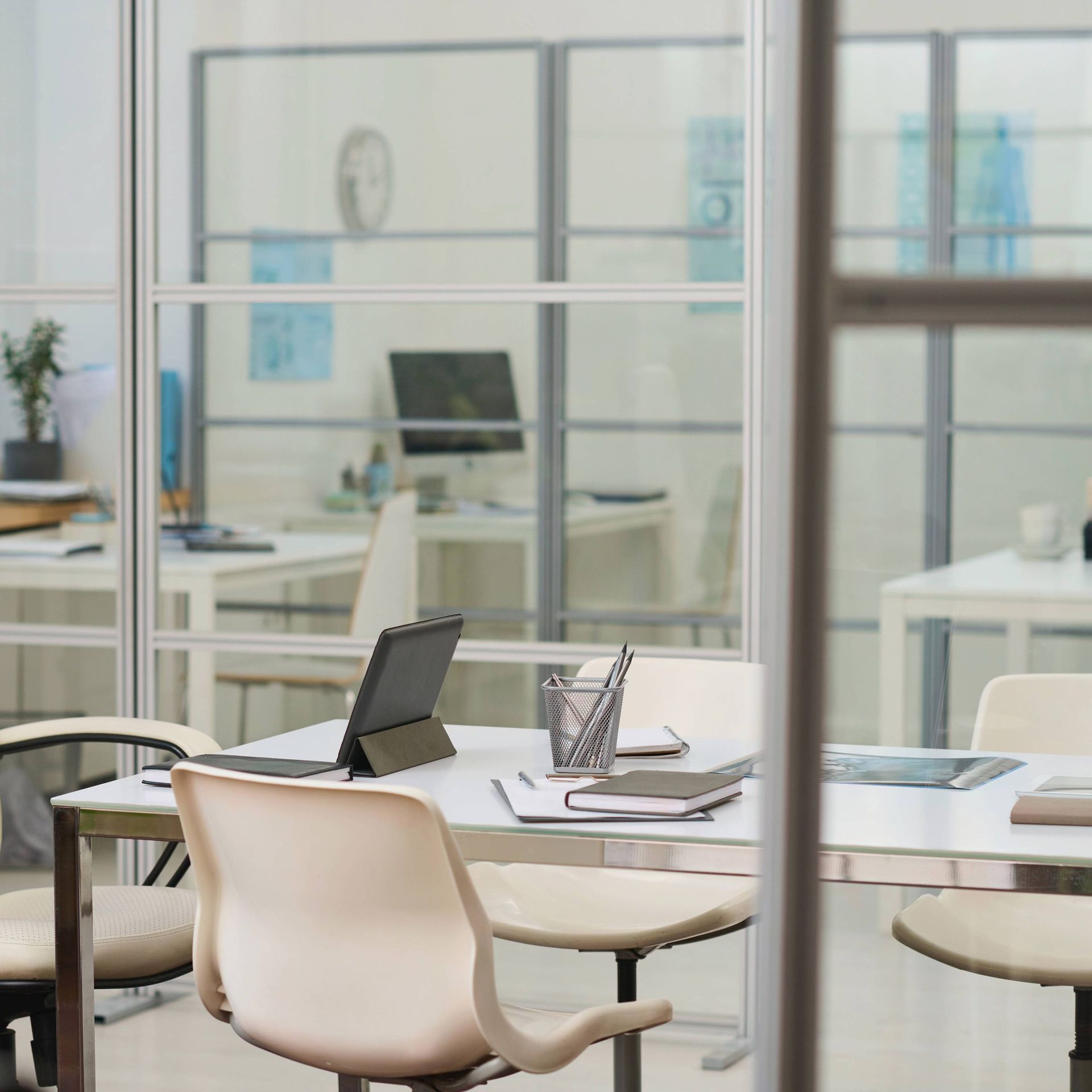 A modern office space with white desks, light-colored chairs, a tablet, and notebooks viewed through glass partitions. A modern office space with white desks, light-colored chairs, a tablet, and notebooks viewed through glass partitions.
