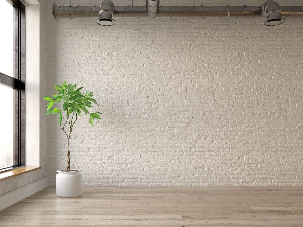 A potted plant sits in a minimalist room with a white brick wall, light wood floor, and industrial exposed ductwork.