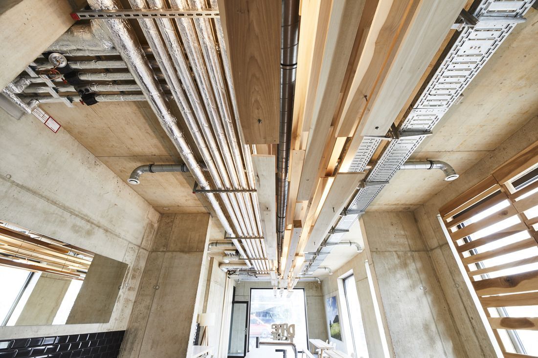 Low-angle view of building utilities, including pipes and a metal cable tray attached to a wooden ceiling above concrete.