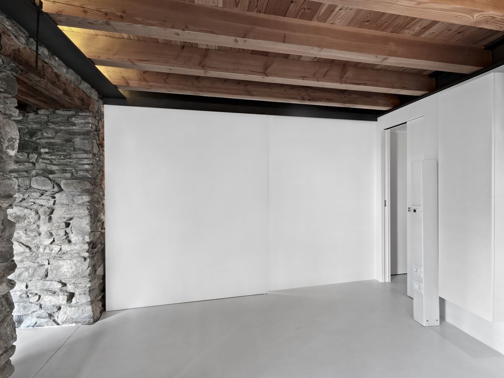A minimalist room with a white wall, a grey floor, exposed wooden ceiling beams, and a natural stone pillar.