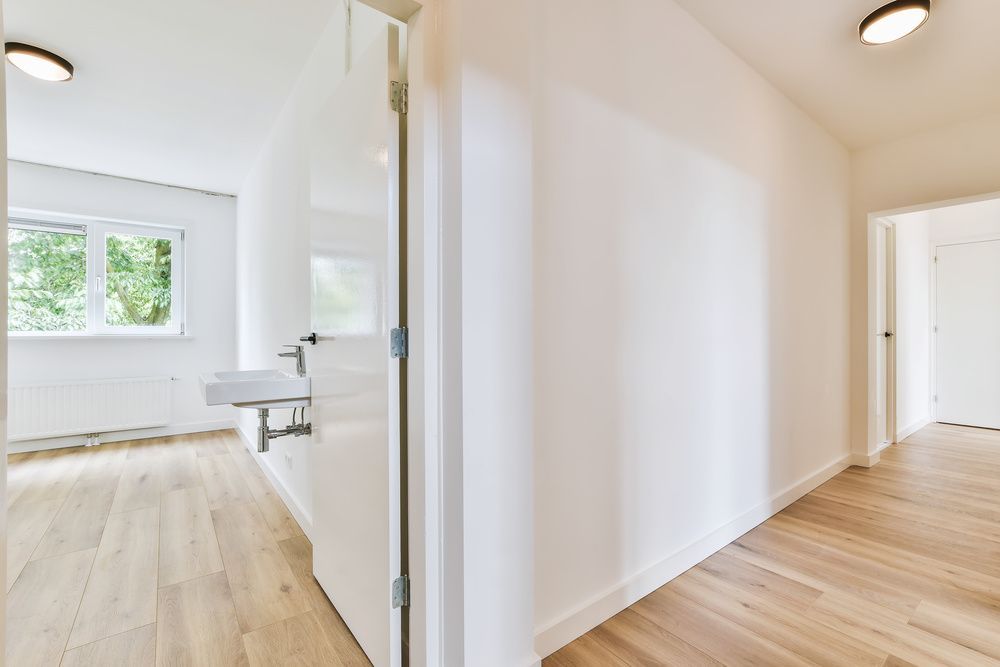 A bright, modern hallway with light wood floors leading to a room with a white wall-mounted sink and large window.