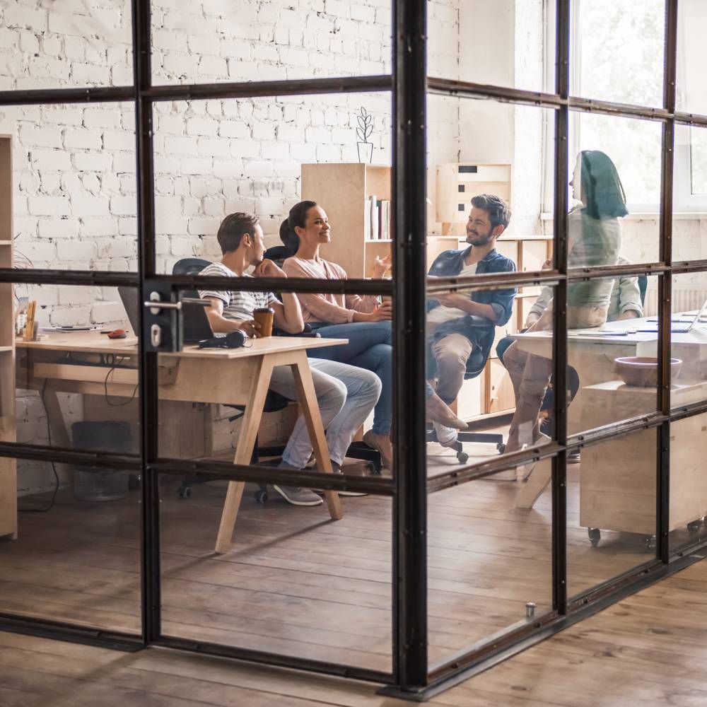 Four people collaborate in a modern, light-filled office with glass walls and white brick, engaged in a group discussion. Four people collaborate in a modern, light-filled office with glass walls and white brick, engaged in a group discussion.