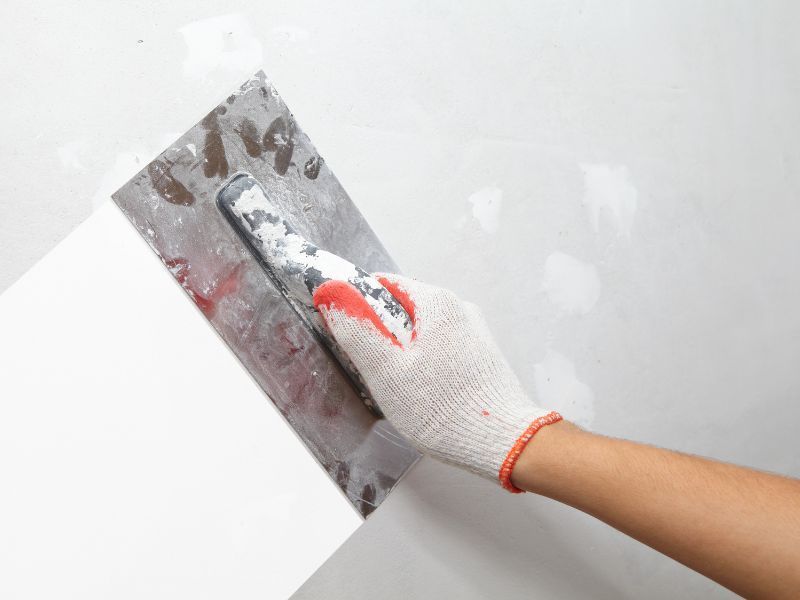 A hand in a white work glove uses a metal trowel to spread joint compound or plaster across a white wall. A hand in a white work glove uses a metal trowel to spread joint compound or plaster across a white wall.