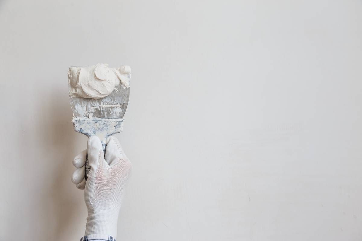 A hand in a white glove holds a putty knife covered in white spackle against a blank white wall. A hand in a white glove holds a putty knife covered in white spackle against a blank white wall.