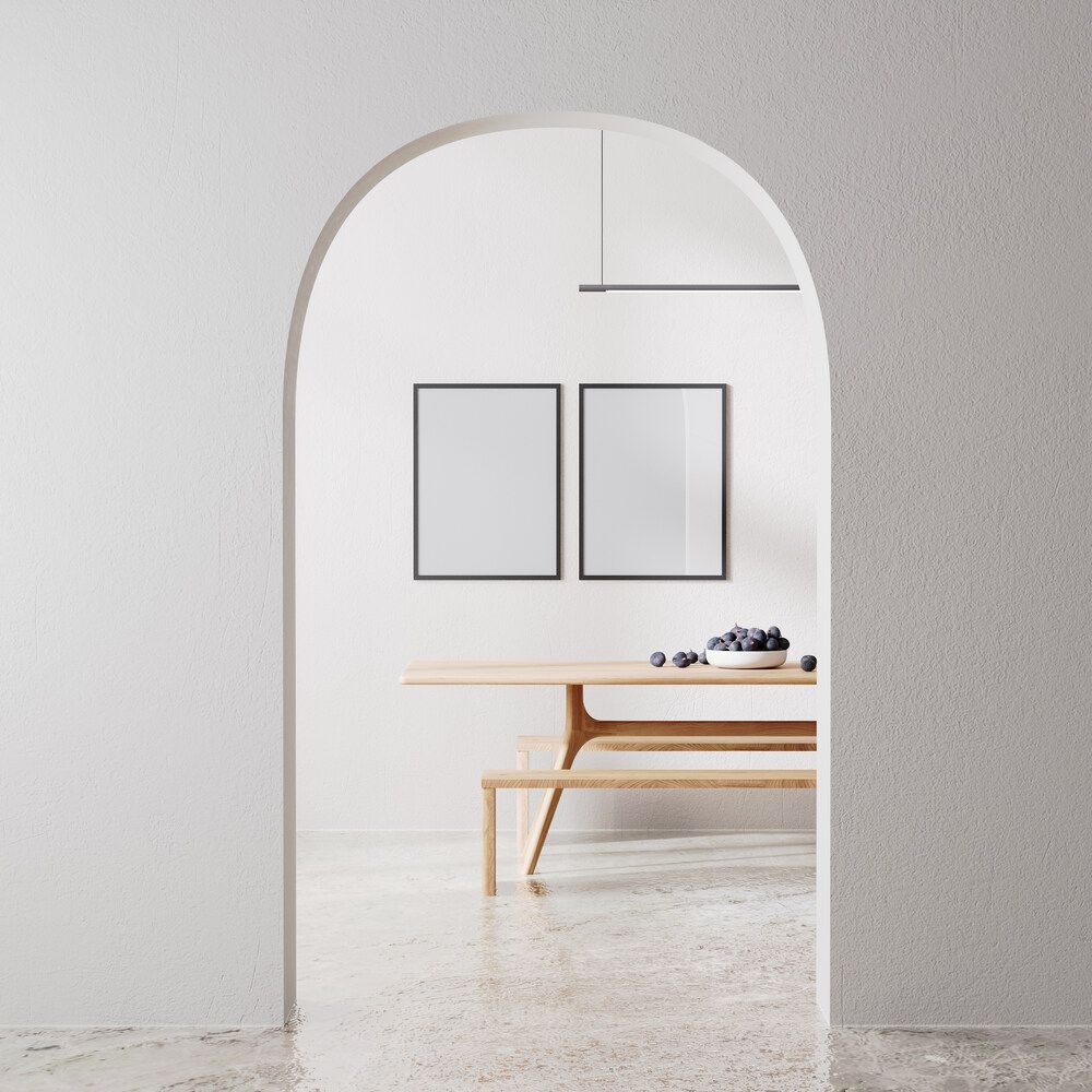 A minimalist dining room viewed through an arched doorway, featuring two blank wall frames above a wooden table and bench.