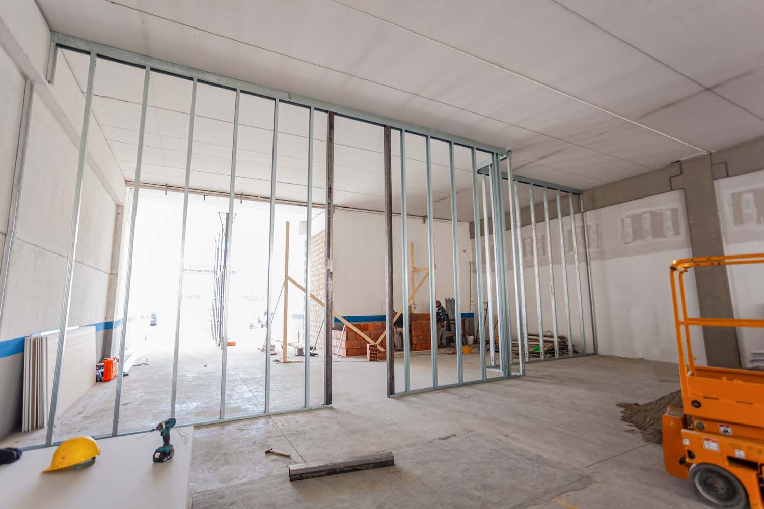 A metal stud frame for a wall being built inside an industrial space, with a yellow hard hat on a desk in the foreground. A metal stud frame for a wall being built inside an industrial space, with a yellow hard hat on a desk in the foreground.