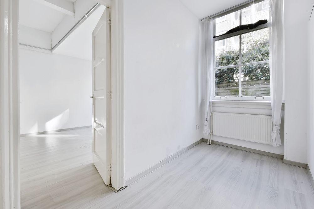 An empty, minimalist room with white walls, light wood-look flooring, a radiator, and a large window with sheer curtains.