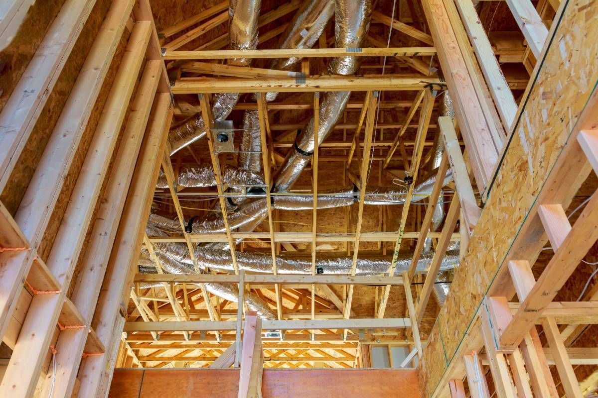 Interior view of wooden wall studs and roof trusses under construction, with silver ventilation ducts visible above.