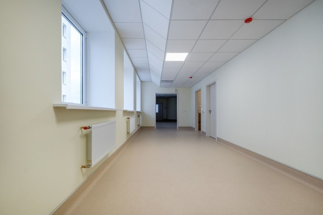 A bright, empty hospital corridor with light-colored walls, large windows on the left, and doors on the right.